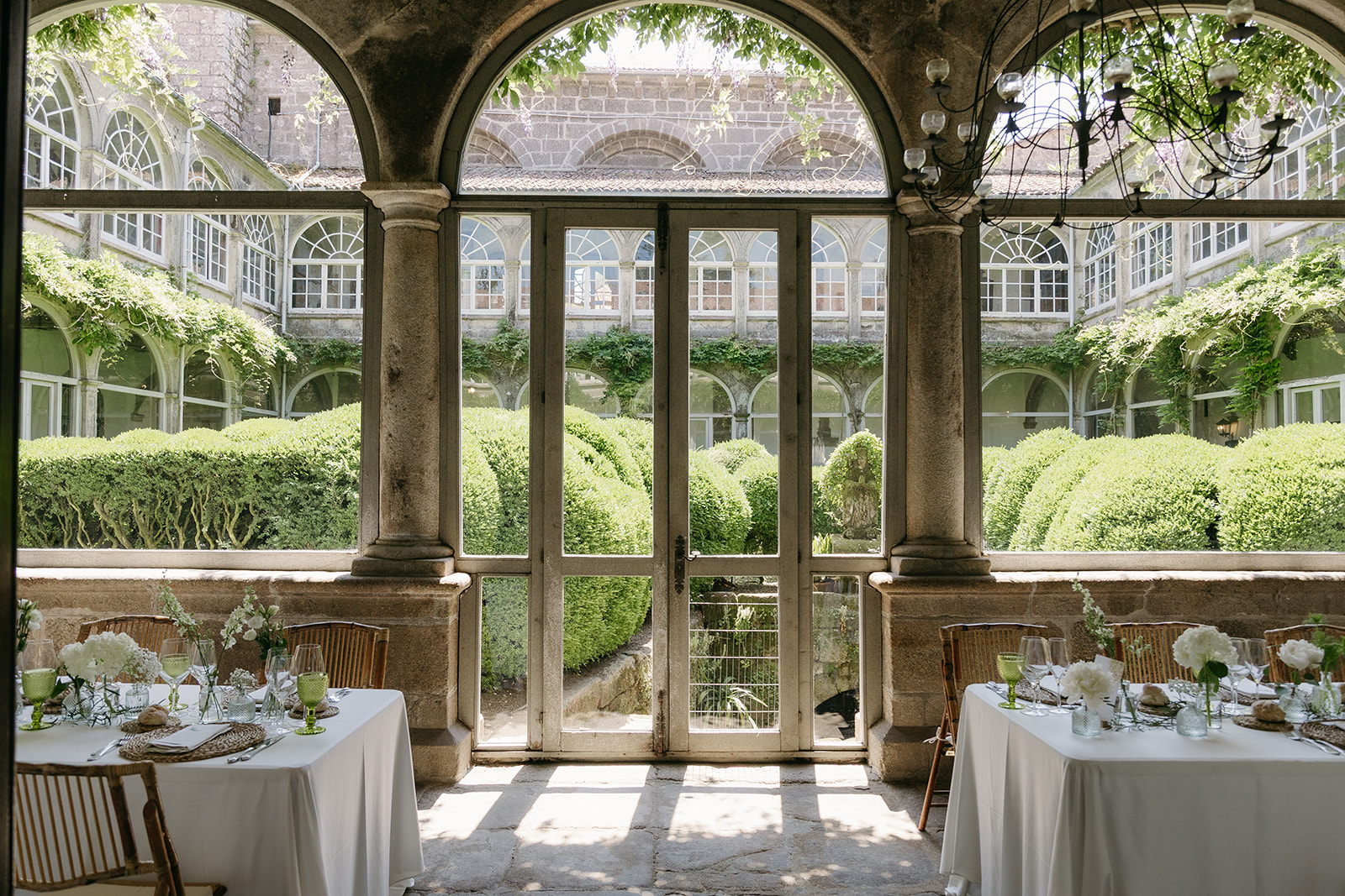 Mesas de boda con flores blancas en un claustro de piedra con vistas a un jardín en Galicia.