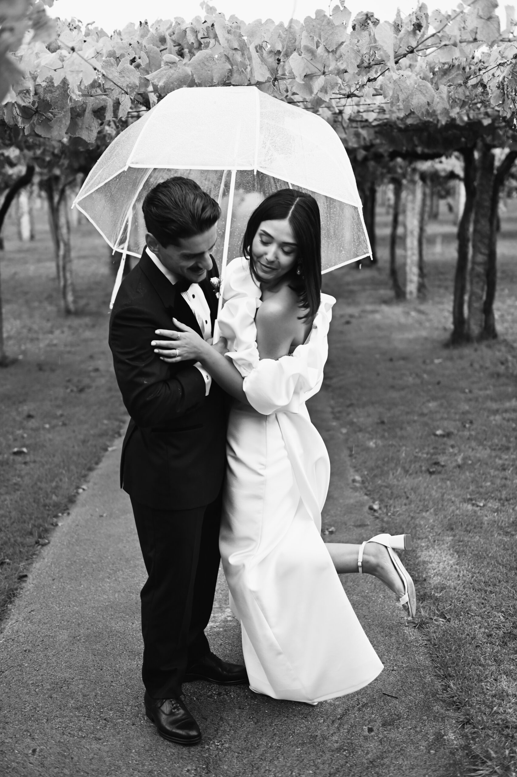 Alejandra y Rubén abrazados bajo un paraguas transparente durante su boda en Galicia, entre los viñedos.