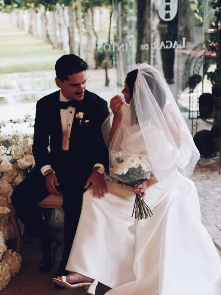 Alejandra & Rubén | Ceremonia de boda en Galicia Alejandra y Rubén sentados durante su ceremonia de boda, rodeados de arreglos florales blancos en un espacio cubierto con vistas al viñedo.