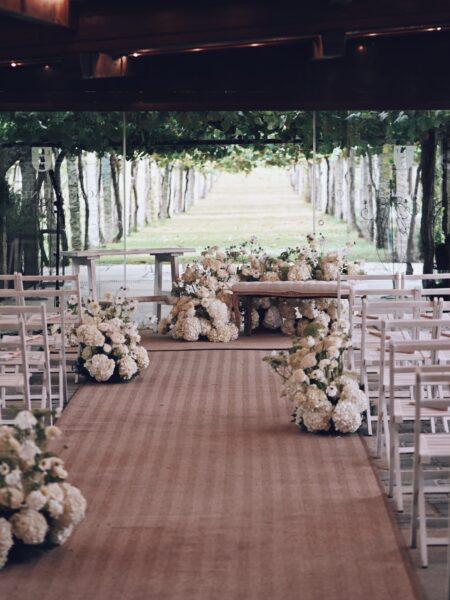 Alejandra & Rubén | Decoración floral de ceremonia en Galicia Bodegas Lagar de Pintos Espacio de ceremonia de boda con pasillo decorado con flores blancas, sillas claras y mesa de ceremonia frente a los viñedos.