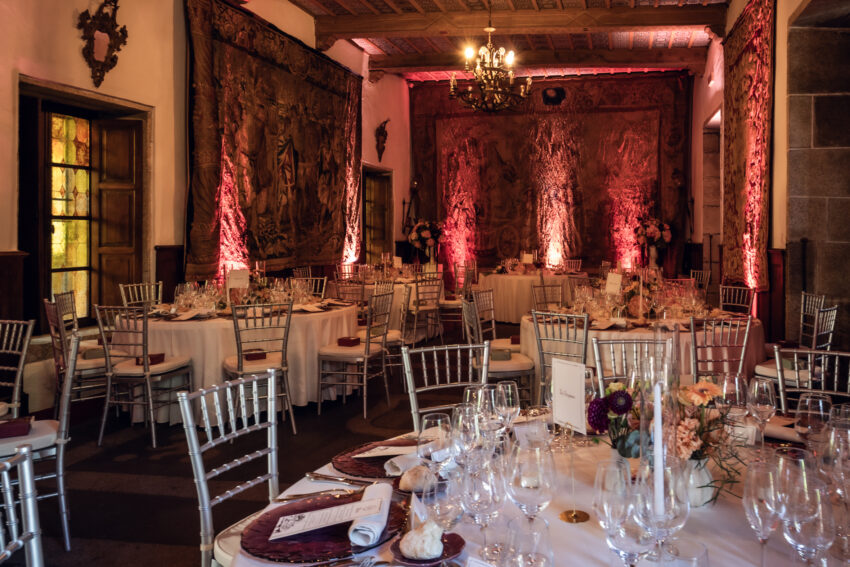 Julia & Aidan | Decoración del salón de bodas en Galicia Salón del banquete de Julia y Aidan con mesas redondas, arreglos florales, sillas plateadas y tapices históricos iluminados en tonos rojizos.