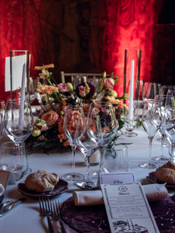 Julia & Aidan | Decoración de mesa y papelería de boda Mesa de boda con flores rosas, velas, cristalería, bajoplatos granates y menú impreso durante el banquete de Julia y Aidan.