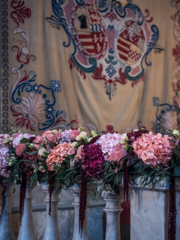 Julia & Aidan | Flores y decoración de iglesia en Galicia Decoración de iglesia con hortensias rosas y granates sobre una balaustrada de piedra, bajo un tapiz histórico.