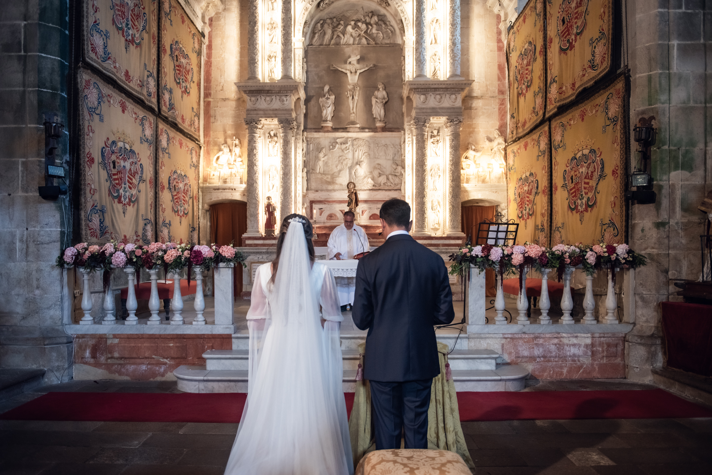 Julia y Aidan de espaldas durante su ceremonia religiosa, frente al altar decorado con flores rosas y granates.